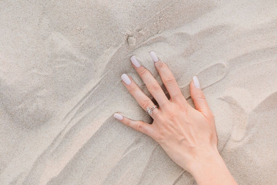 Primer plano de una mano femenina con un anillo de diamantes y uñas bien cuidadas sobre arena.