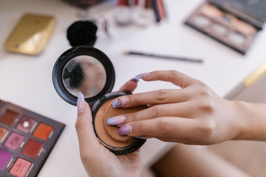 Manos aplicando polvo con una esponja de un compacto, rodeado de productos de maquillaje.