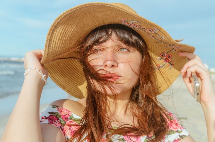 Mujer joven en la playa con sombrero de paja y top floral disfrutando de un día soleado.