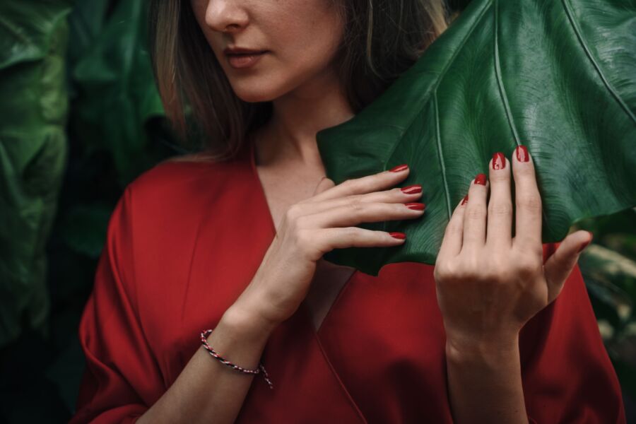 Una mujer con un elegante atuendo rojo tocando suavemente una gran hoja verde. Ambiente elegante y natural.