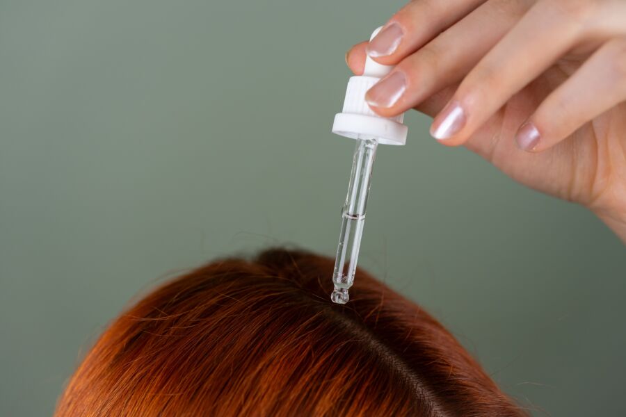 Primer plano de la aplicación de suero a las raíces con un cuentagotas de precisión sobre el cabello rojo, mostrando la rutina de cuidado del cabello.