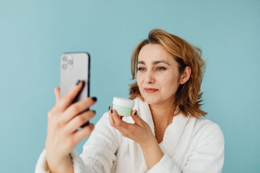 Mujer en bata blanca tomándose una selfie mientras sostiene crema para el cuidado de la piel sobre un fondo azul.