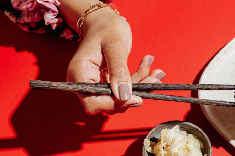 Una mano de mujer con uñas bien cuidadas sosteniendo palillos sobre un fondo rojo, mostrando elegancia.