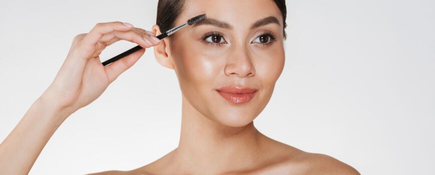 Ciérrese encima del retrato del estudio de la mujer morena hermosa con la piel suave que pinta sus cejas con el cepillo del maquillaje, aislado sobre blanco