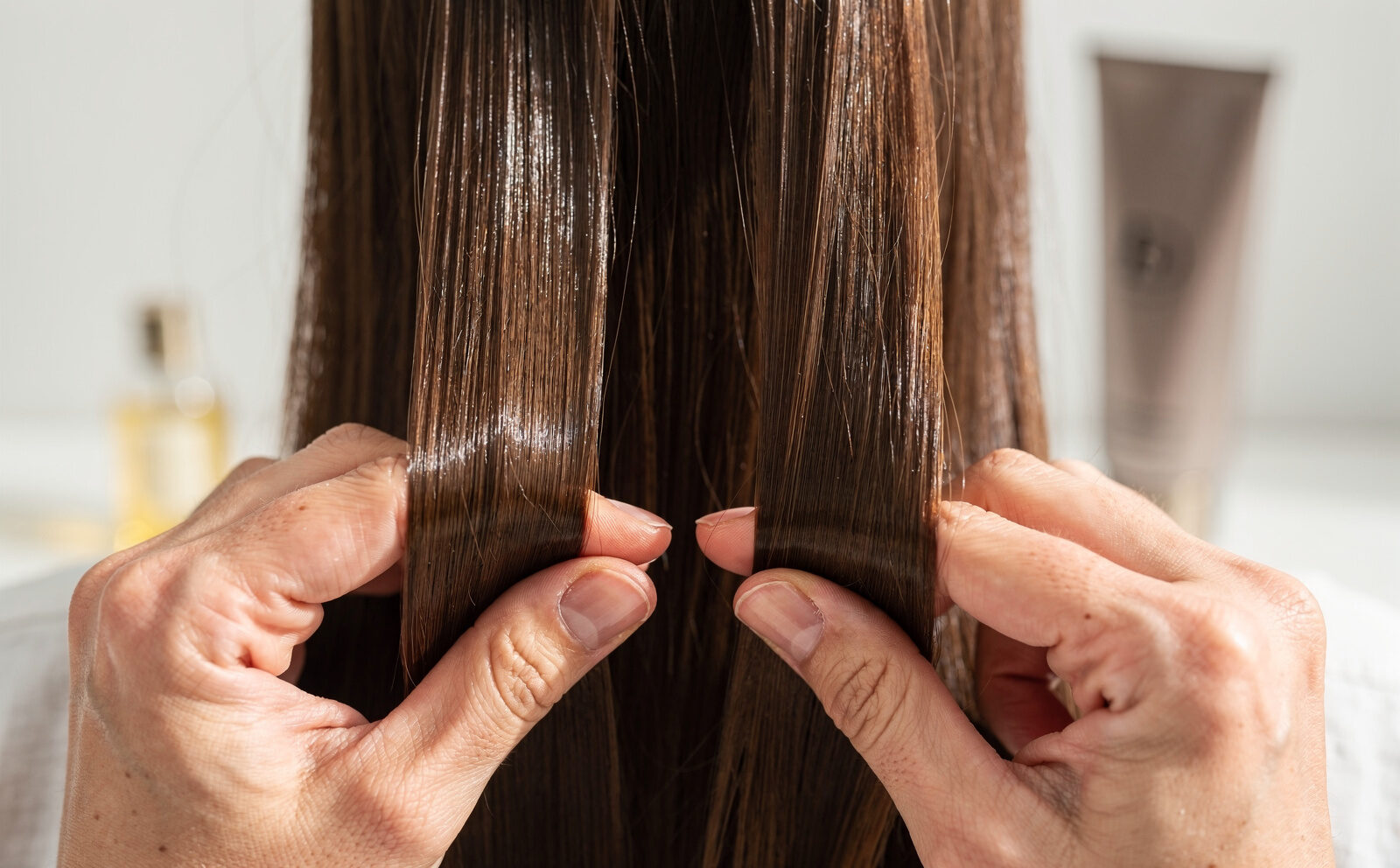 Manos aplicando en dos mechones de pelo dos productos distintos como para hacer un contraste de resultados. Se intuyen los envases de Olaplex Nº7 Bonding Oil y Kérastase Nutritive de fondo a cada lado del mechón