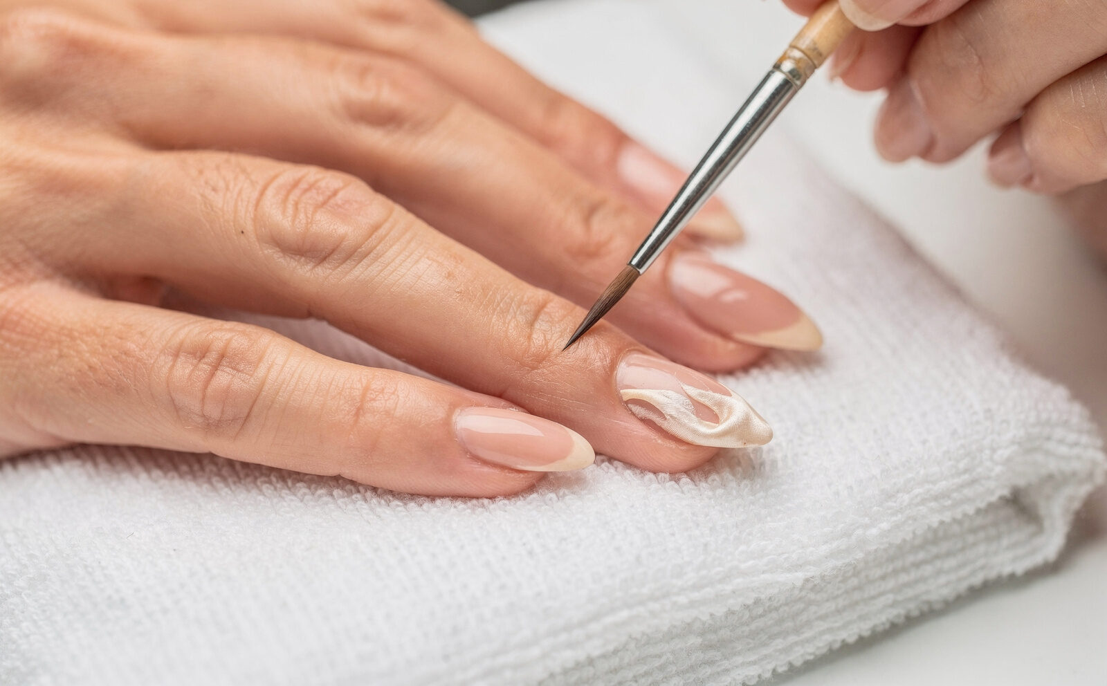Pincel fino pintando un diseño satinado tipo seda sobre una uña almendrada, mano apoyada en toalla de manicura; proceso en curso, sin mano terminada.