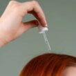 Primer plano de la mano aplicando suero capilar sobre el cabello rojo con un gotero.