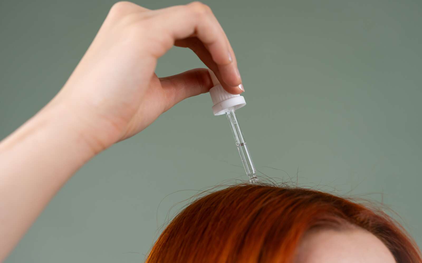 Primer plano de la mano aplicando suero capilar sobre el cabello rojo con un gotero.