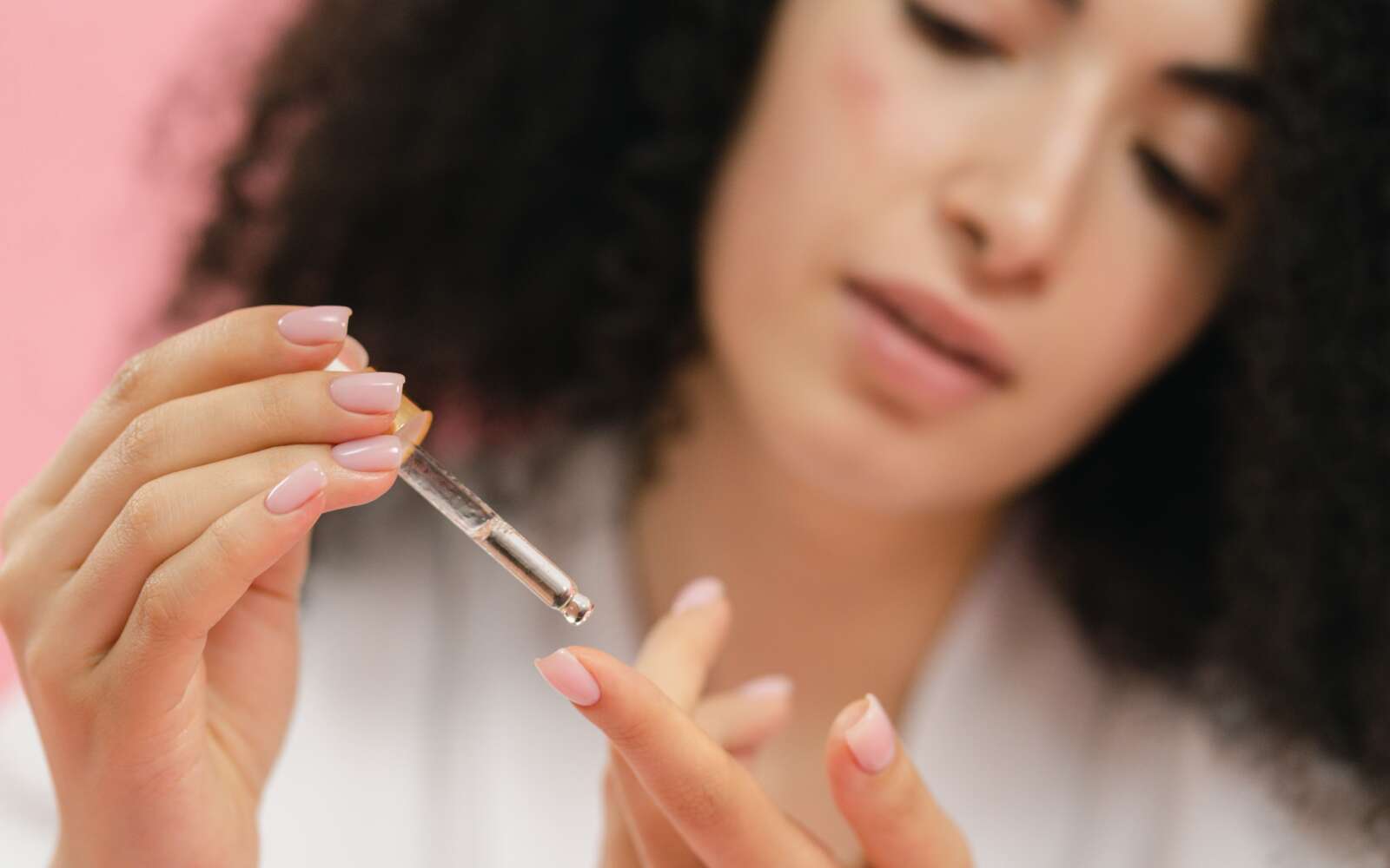 Primer plano de una mujer joven aplicando suero para el cuidado de la piel con una pipeta sobre un fondo rosa.
