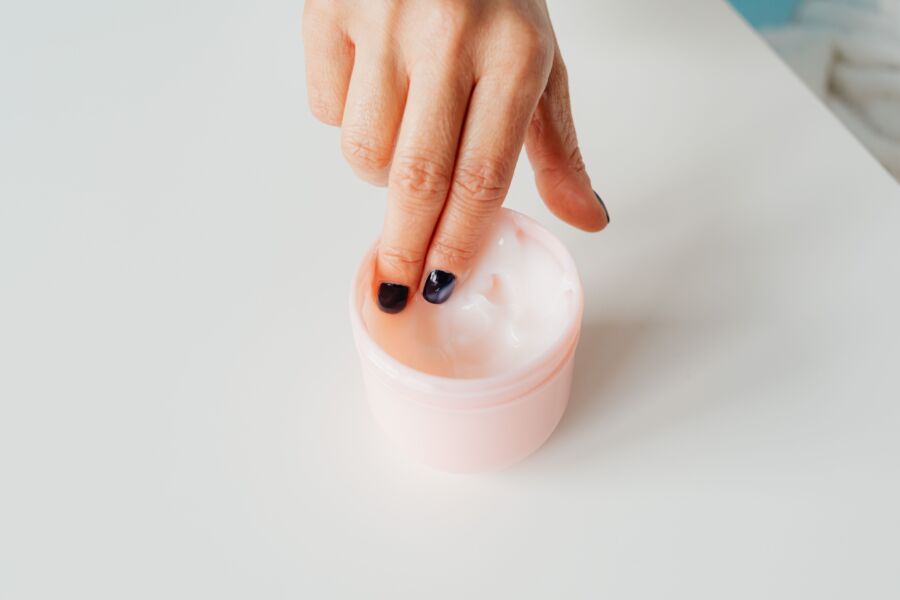 Primer plano de una mano con las uñas pintadas que se sumerge en un frasco de crema facial, que representa el autocuidado.