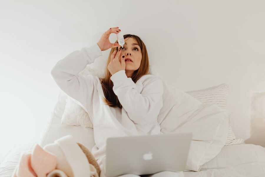 Una mujer aplica gotas para los ojos mientras descansa en la cama con un ordenador portátil, que simboliza la relajación y el autocuidado en casa.