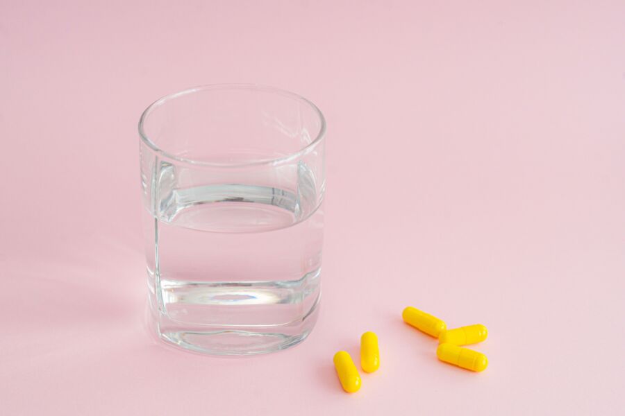 Un vaso de agua transparente junto a cápsulas amarillas sobre un fondo rosa, que simboliza el cuidado de la salud.