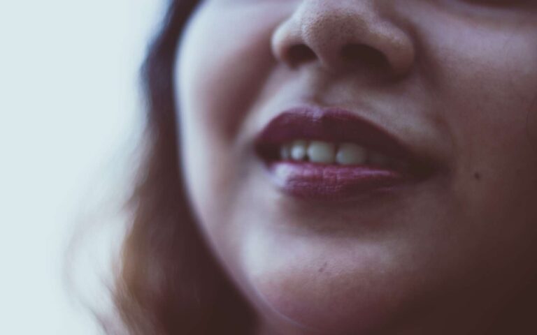 Primer plano de la cara de una mujer alegre irreconocible con cabello suelto oscuro con lápiz labial brillante y sonrisa dentada sobre fondo borroso a la luz del día