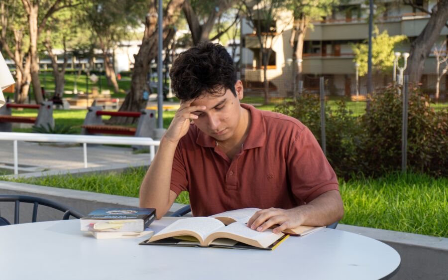 Un joven que se concentra intensamente mientras estudia en un entorno universitario parecido a un parque en Piura, Perú.