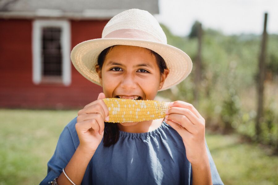 Alegre niña hispana con sombrero que muerde un sabroso y apetitoso maíz mientras pasa el día de verano en el patio