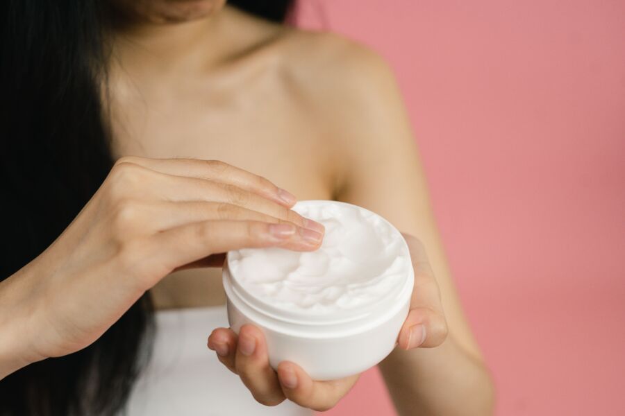 Primer plano de una mujer aplicando crema blanca de un frasco sobre un fondo rosado.