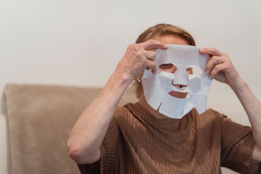 Mujer mayor aplicando una mascarilla de sábanas para el cuidado de la piel y la relajación en interiores.