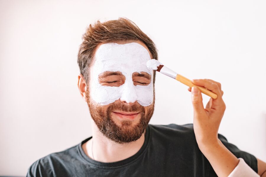 Un hombre con barba disfruta de un tratamiento de spa en casa con una mascarilla aplicada con un cepillo.