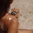 Mujer que se aplica protector solar en el hombro en un día soleado de playa, centrándose en el cuidado de la piel.
