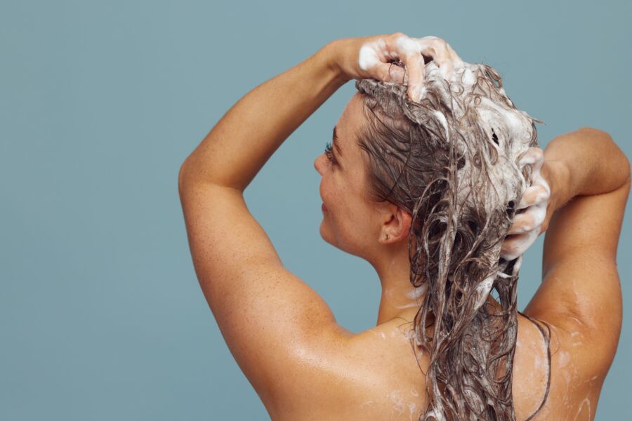 Mujer con cabello largo lavándolo con champú, creando espuma, en ambiente interior sobre fondo azul.