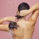Mujer lavándose el pelo largo bajo la ducha con gotas de agua sobre un fondo rosado.