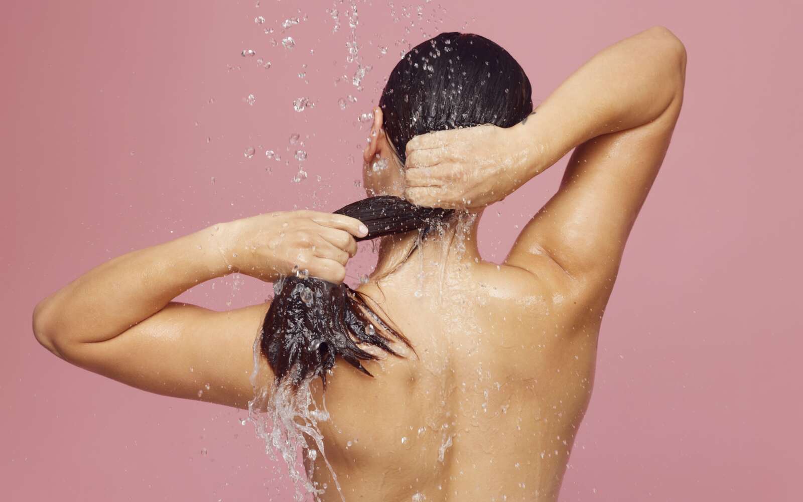Mujer lavándose el pelo largo bajo la ducha con gotas de agua sobre un fondo rosado.