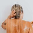 Vista posterior de una mujer enjuagándose el cabello bajo la ducha, capturando un momento íntimo y refrescante en el interior.