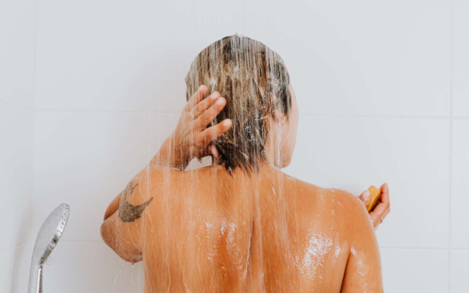 Vista posterior de una mujer enjuagándose el cabello bajo la ducha, capturando un momento íntimo y refrescante en el interior.