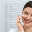 Mujer sonriente en bata blanca con crema facial en el baño.