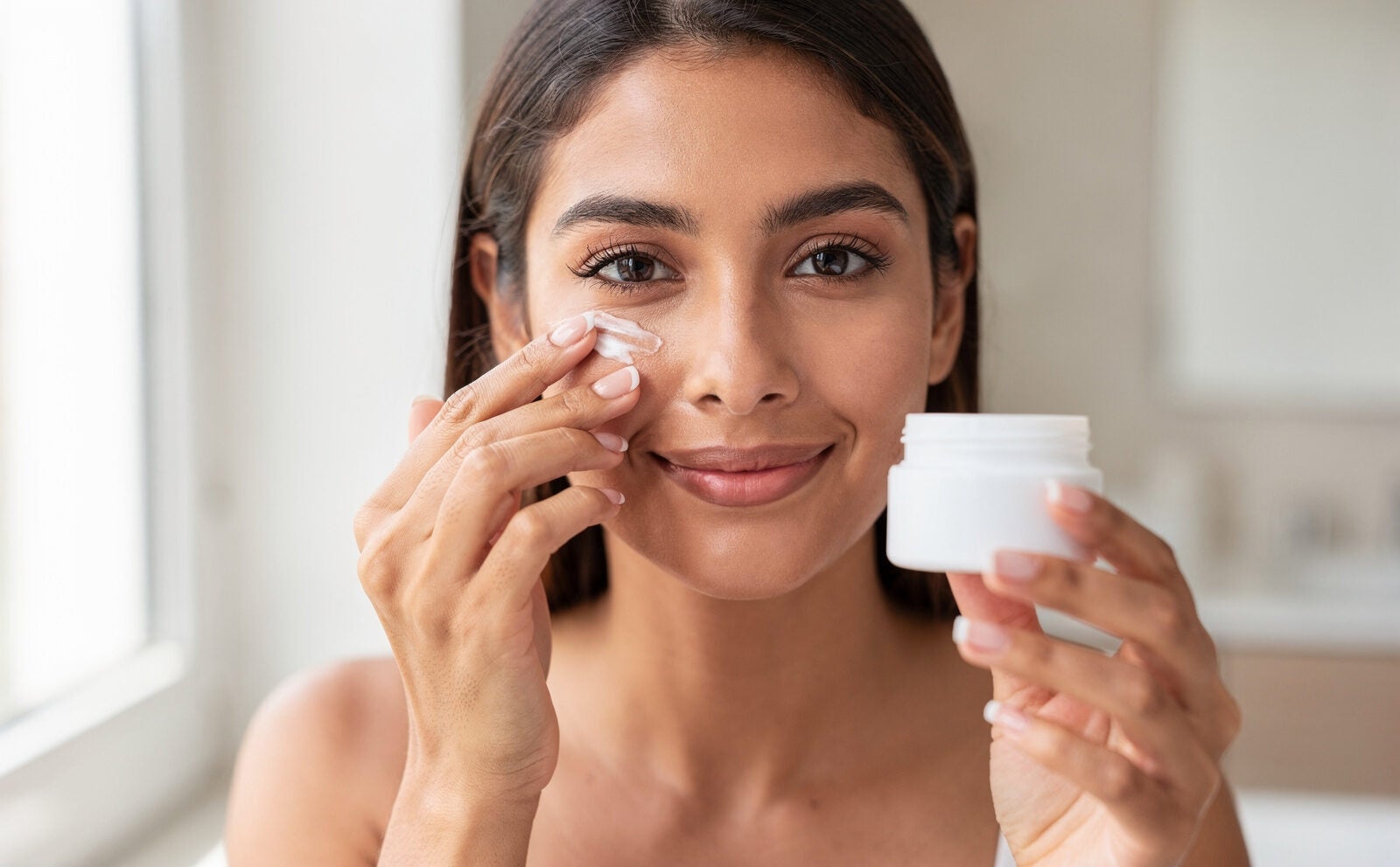 chica se pone crema en el contorno de los ojos y tiene en la mano el bote de crema