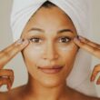 Mujer aplicando crema facial con una toalla envuelta en la cabeza, destacando rutina de cuidado de la piel.