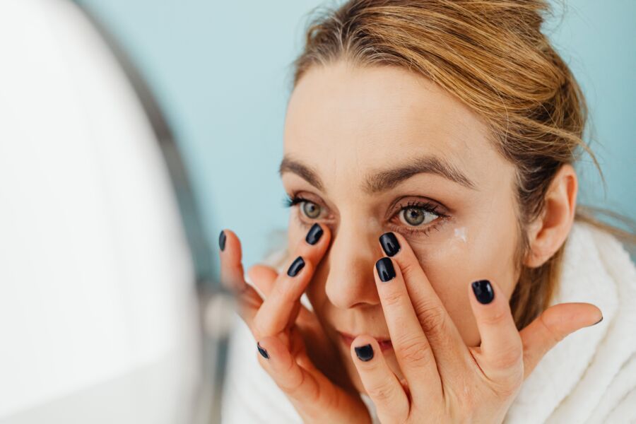 Primer plano de una mujer aplicando crema facial frente a un espejo con fondo azul.