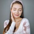 Retrato de una mujer sonriente con crema facial, los ojos cerrados y la mano en la mejilla sobre un fondo gris.