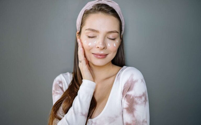 Retrato de una mujer sonriente con crema facial, los ojos cerrados y la mano en la mejilla sobre un fondo gris.