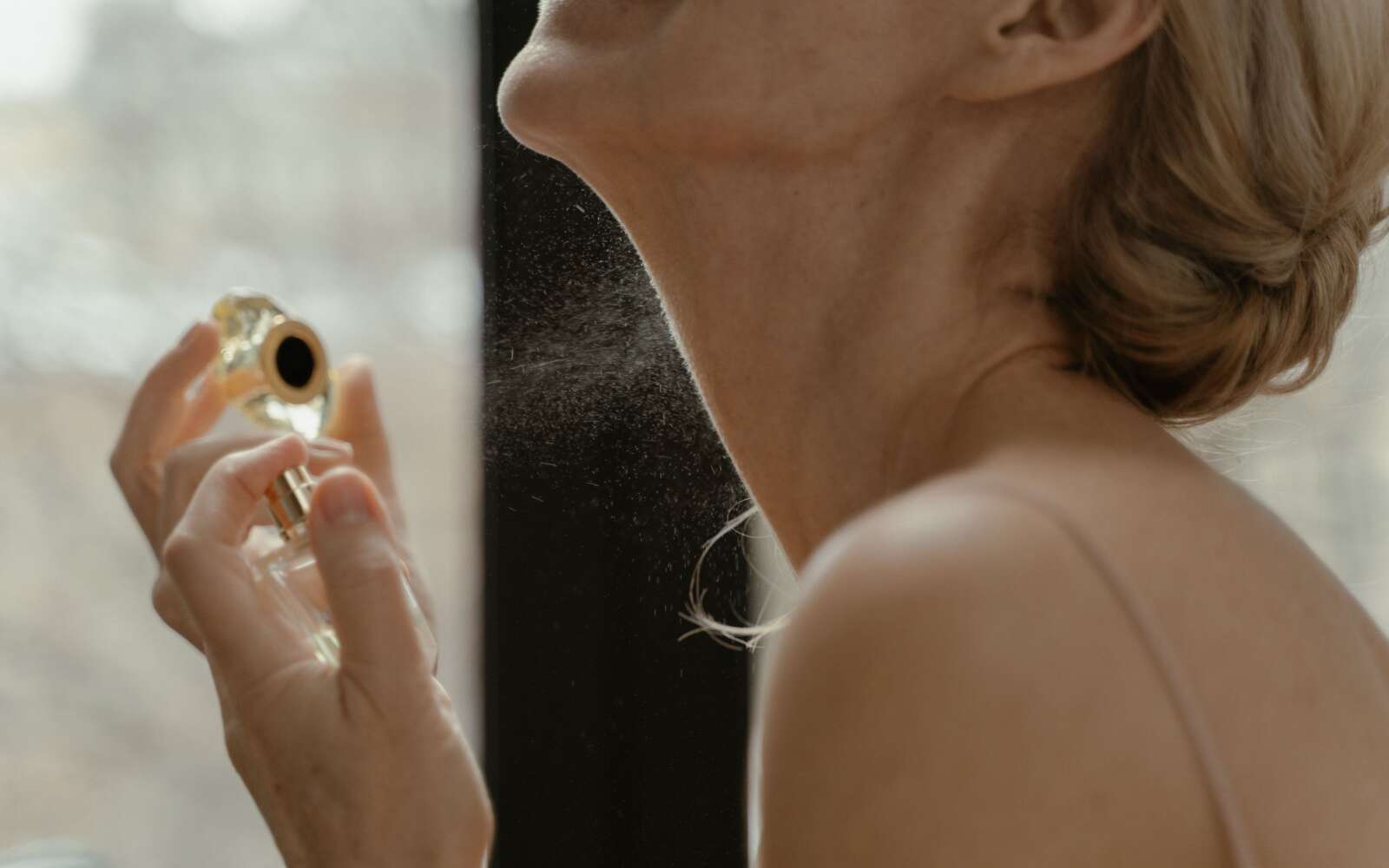 Primer plano de una mujer aplicando perfume con un frasco de vidrio con luz natural.