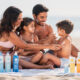 una familia en la playa donde se vea en la toalla todo un surtido de protectores solares de la marca nivea. importante: los productos se ven en segundo plano difumiado, primero la familia poniendose crema