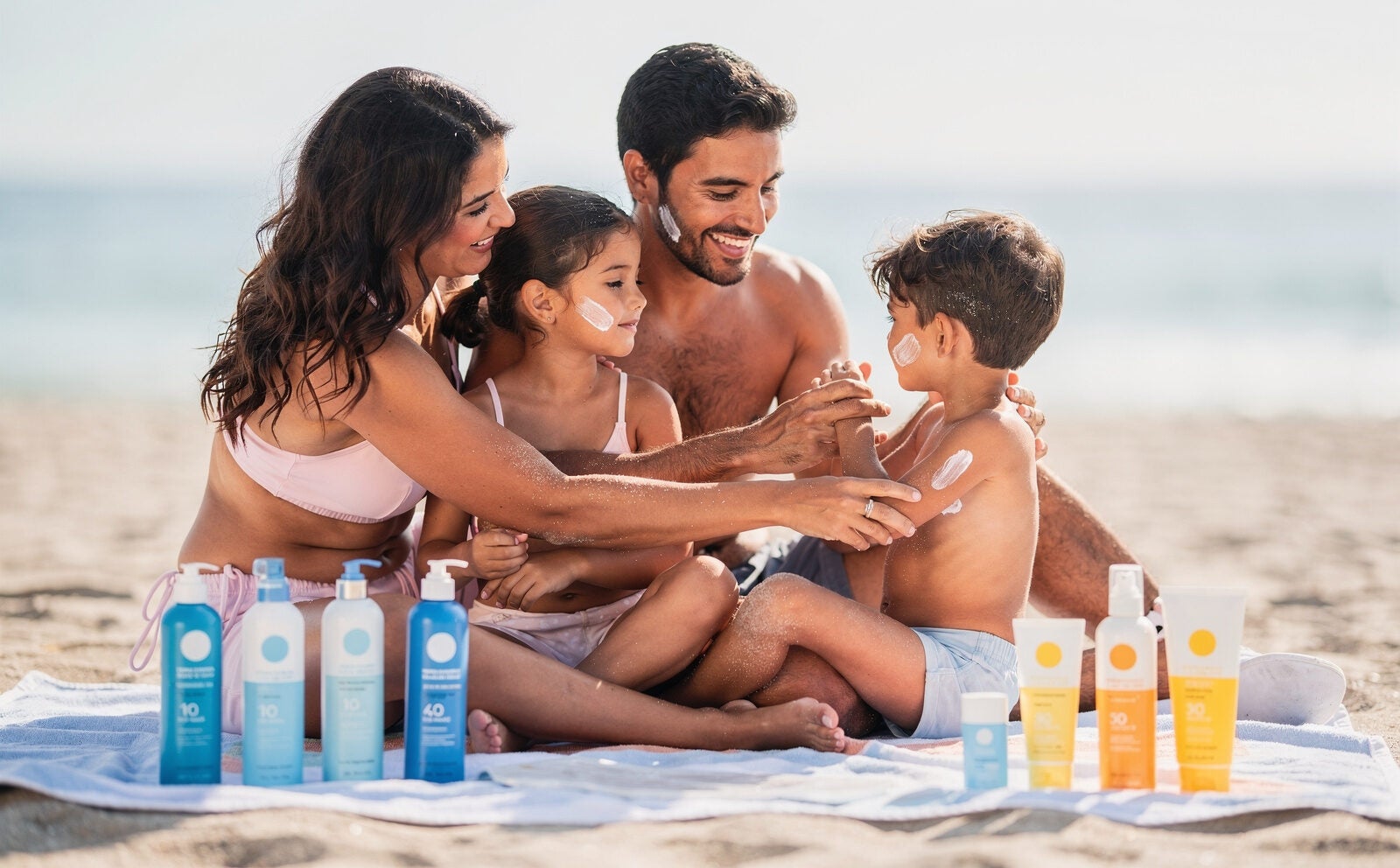una familia en la playa donde se vea en la toalla todo un surtido de protectores solares de la marca nivea. importante: los productos se ven en segundo plano difumiado, primero la familia poniendose crema