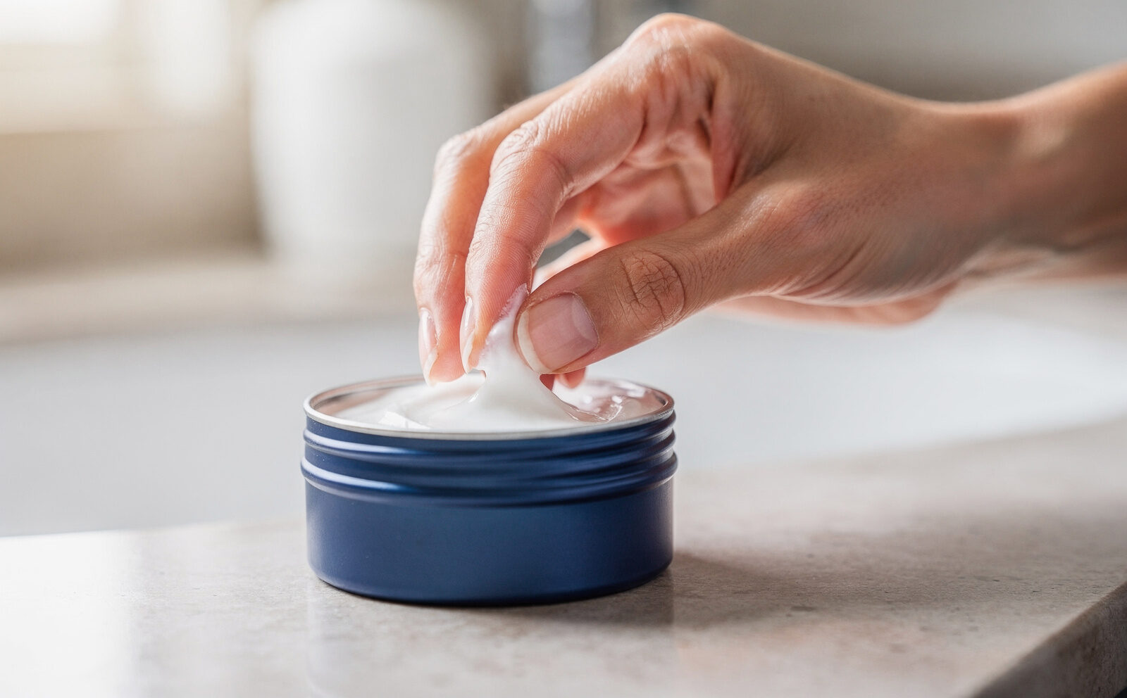 Primer plano de unas manos tomando crema con los dedos directamente de una lata azul abierta, sobre lavabo claro con luz natural. Gesto en curso, sin rostro de persona famosa, sin antes/después.