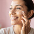 mujer se pone crema facial, fondo borroso, mujer sonriente, tono rosado y limpio