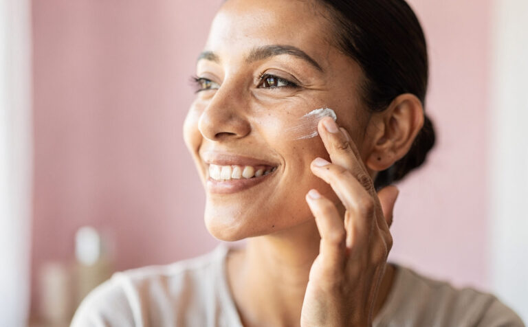 mujer se pone crema facial, fondo borroso, mujer sonriente, tono rosado y limpio