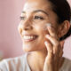 mujer se pone crema facial, fondo borroso, mujer sonriente, tono rosado y limpio