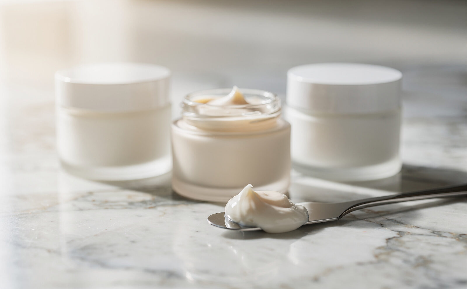 Bodegón vertical de 3 tarros de crema facial sin etiquetas visibles sobre mármol claro, con una gota de textura en espátula cosmética, luz natural lateral. Sin rostros ni comparativas antes/después.