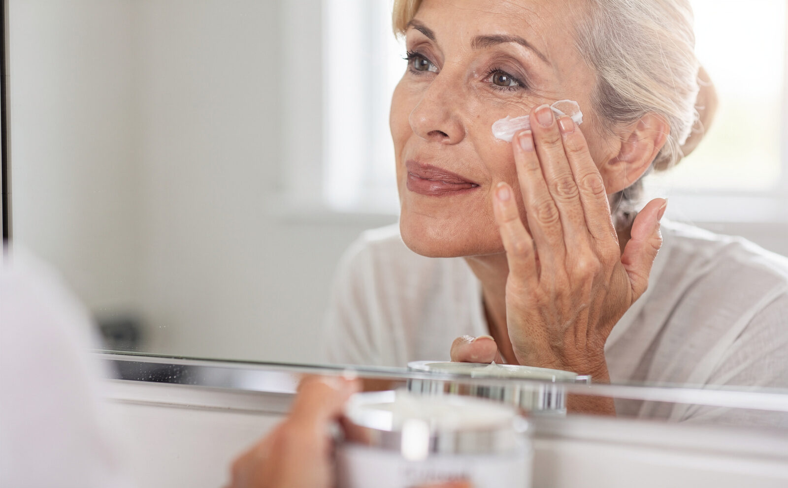 Mujer de unos 55 años aplicándose crema con las yemas de los dedos en el pómulo, frente al espejo del baño con luz natural; tarro abierto en primer plano. Gesto en curso, sin antes/después.