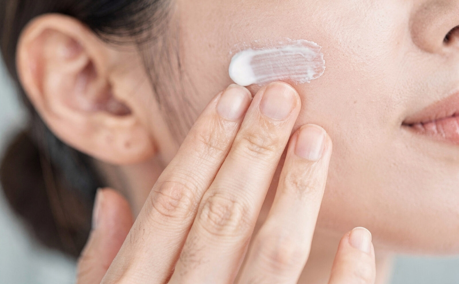 Manos aplicando la crema con gesto ascendente sobre el pómulo, foco en el proceso. Regla anti-resultado: no mostrar el rostro ya con la crema absorbida ni comparación antes/después. Sin celebridades.