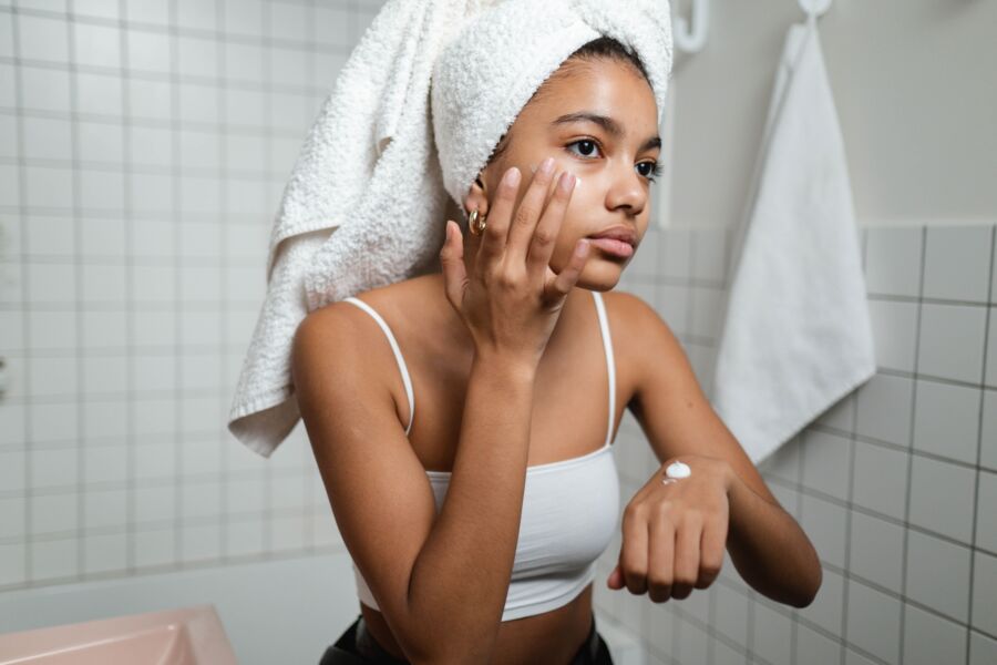 Una mujer joven en un baño aplicando crema para el cuidado de la piel con una toalla en la cabeza, centrándose en el autocuidado.