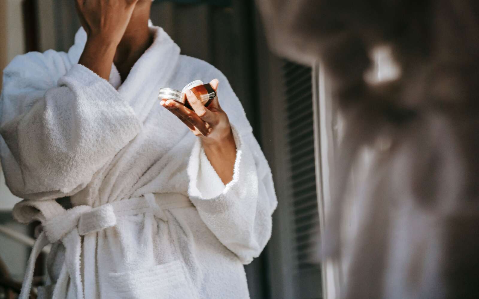 Recorte mujer étnica anónima en suave bata de baño aplicando crema nutritiva en la cara mientras se refleja en el espejo en casa