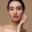 Mujer sensible al recorte con maquillaje y labios rojos aplicando crema sobre la delicada piel de la cara mientras mira la cámara sobre fondo beige