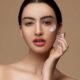 Mujer sensible al recorte con maquillaje y labios rojos aplicando crema sobre la delicada piel de la cara mientras mira la cámara sobre fondo beige
