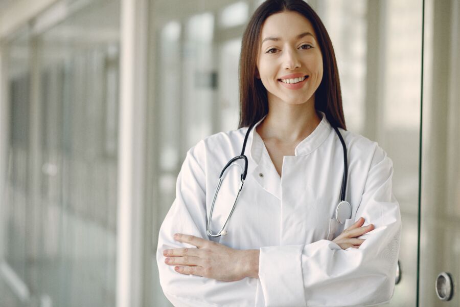 Recortar alegre doctora étnica en uniforme médico y estetoscopio con las manos cruzadas de pie en el moderno pasillo de la clínica con pared de vidrio
