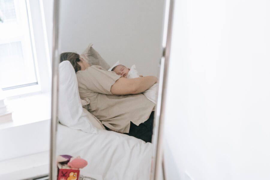 Mujer anónima con bebé dormido en brazos que se refleja en el espejo mientras descansa en una cama cómoda en un dormitorio ligero en casa